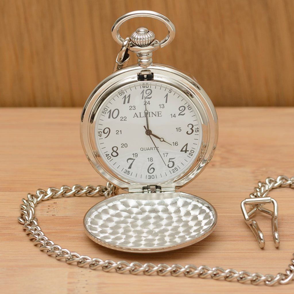 A silver pocket watch with its cover open, resting on a light wood surface. The watch has a silver-tone chain, a hammered-texture lower cover, and a white dial with black numerals.