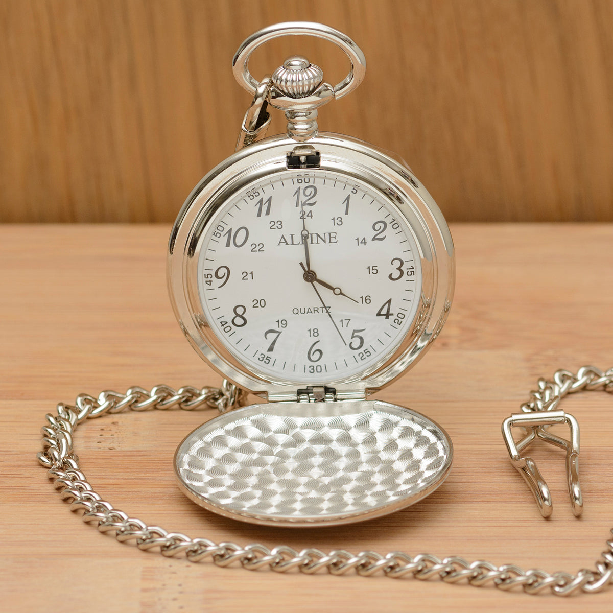 A silver pocket watch with its cover open, resting on a light wood surface. The watch has a silver-tone chain, a hammered-texture lower cover, and a white dial with black numerals.
