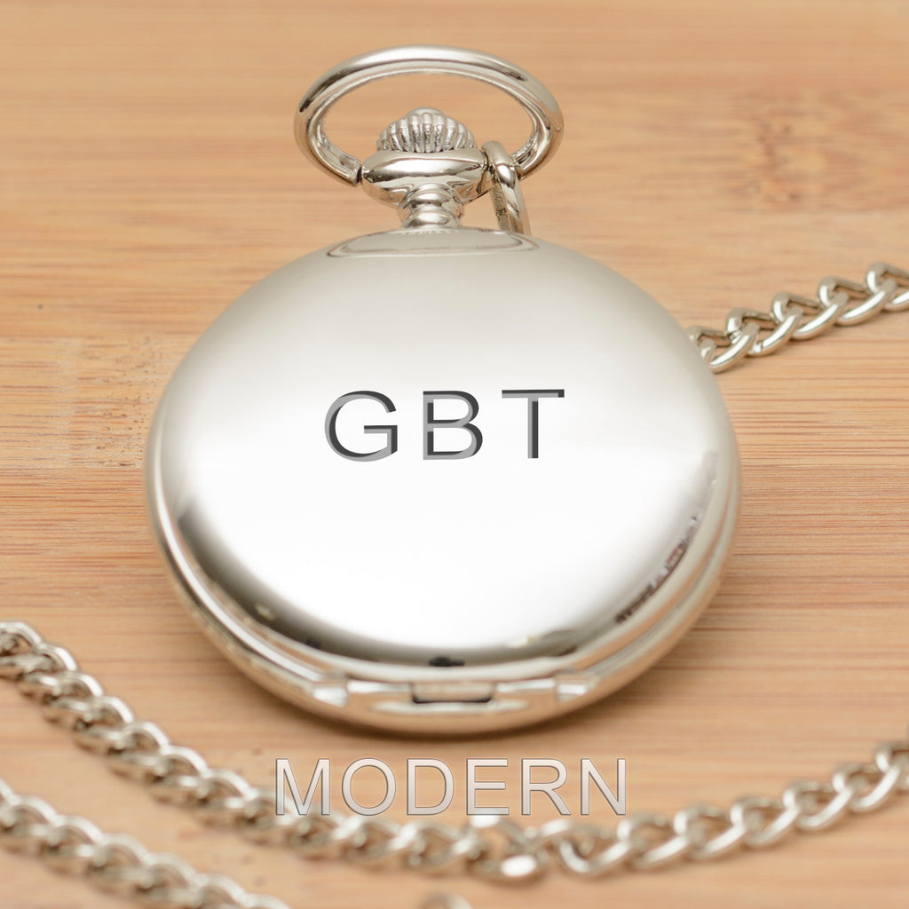 A silver pocket watch pendant with a matching chain, engraved with the initials 'GBT' displayed on a light wood background.
