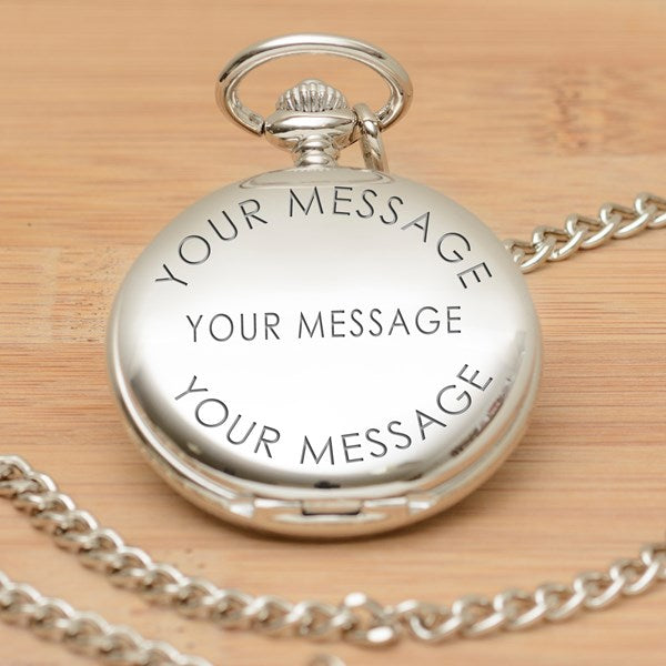 A silver pocket watch pendant with a matching chain, engraved with the wording 'YOUR MESSAGE' at the top, middle and bottom displayed on a light wood background.
