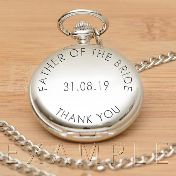 A silver pocket watch pendant necklace with a chain, shown on a light wood background. The back of the round watch is engraved with the words 'FATHER OF THE BRIDE', and the date '31.08.19'.