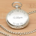 A silver pocket watch pendant with a matching chain, engraved with the wording 'DON'T BE LATE ... 12.00pm 22.08.26' displayed on a light wood background.
