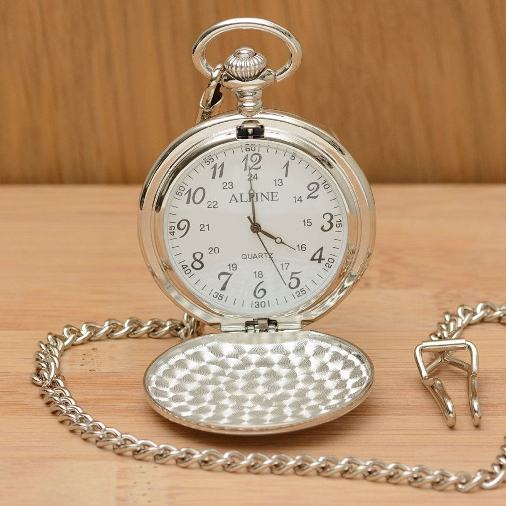 A silver pocket watch with its cover open, resting on a light wood surface. The watch has a silver-tone chain, a hammered-texture lower cover, and a white dial with black numerals.