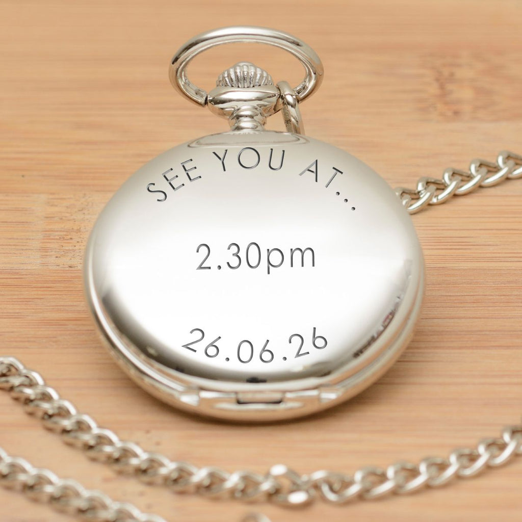 A silver pocket watch pendant with a matching chain, engraved with the wording 'SEE YOU AT ... 2.30pm 26.06.26' displayed on a light wood background.
