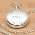 A silver pocket watch pendant with a matching chain, engraved with the wording 'SEE YOU AT ... 2.30pm 26.06.26' displayed on a light wood background.
