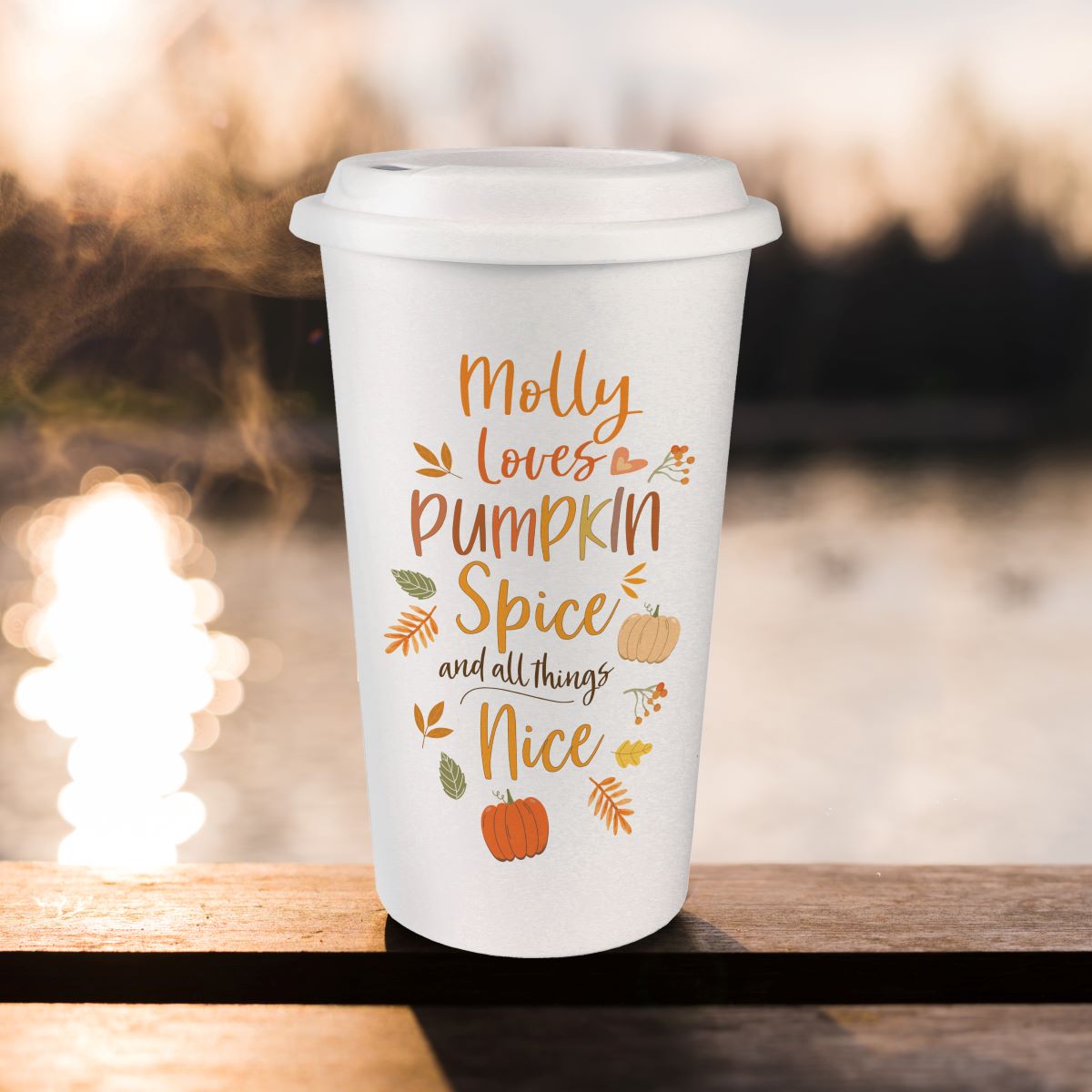 A white reusable coffee cup with a lid sits on a wooden railing outdoors. The cup has a printed autumn-themed quote reading “Molly loves Pumpkin Spice and all things Nice” in orange and brown lettering, with small pumpkins, leaves, and fall motifs.