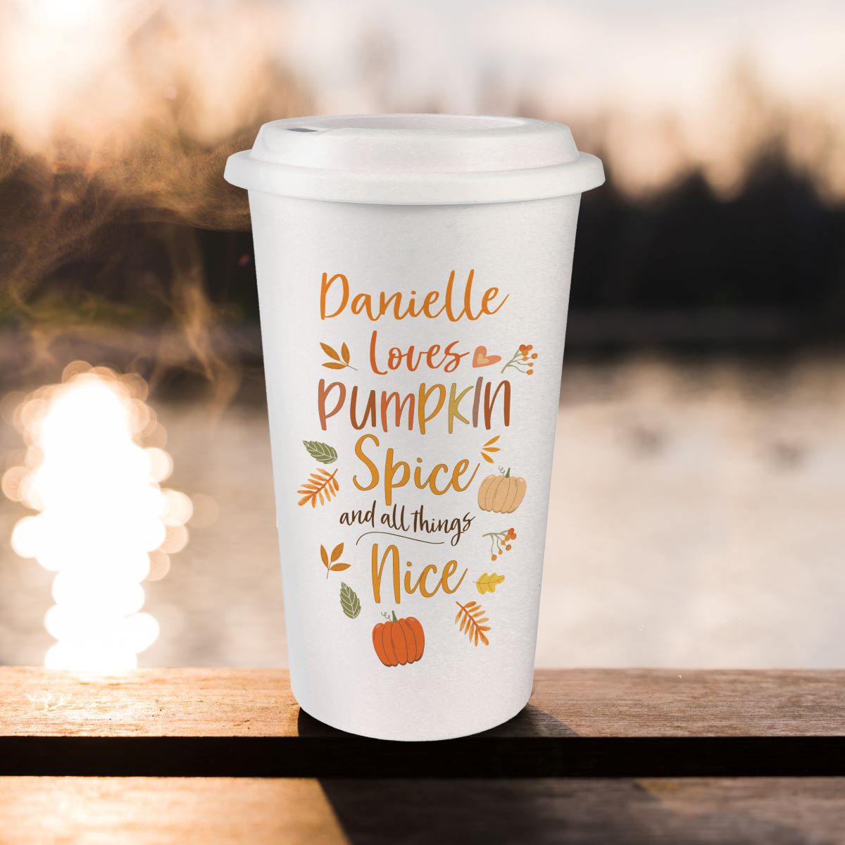 White travel mug with 'Danielle loves Pumpkin Spice and all things Nice' text on a wooden surface with blurred lights in the background.