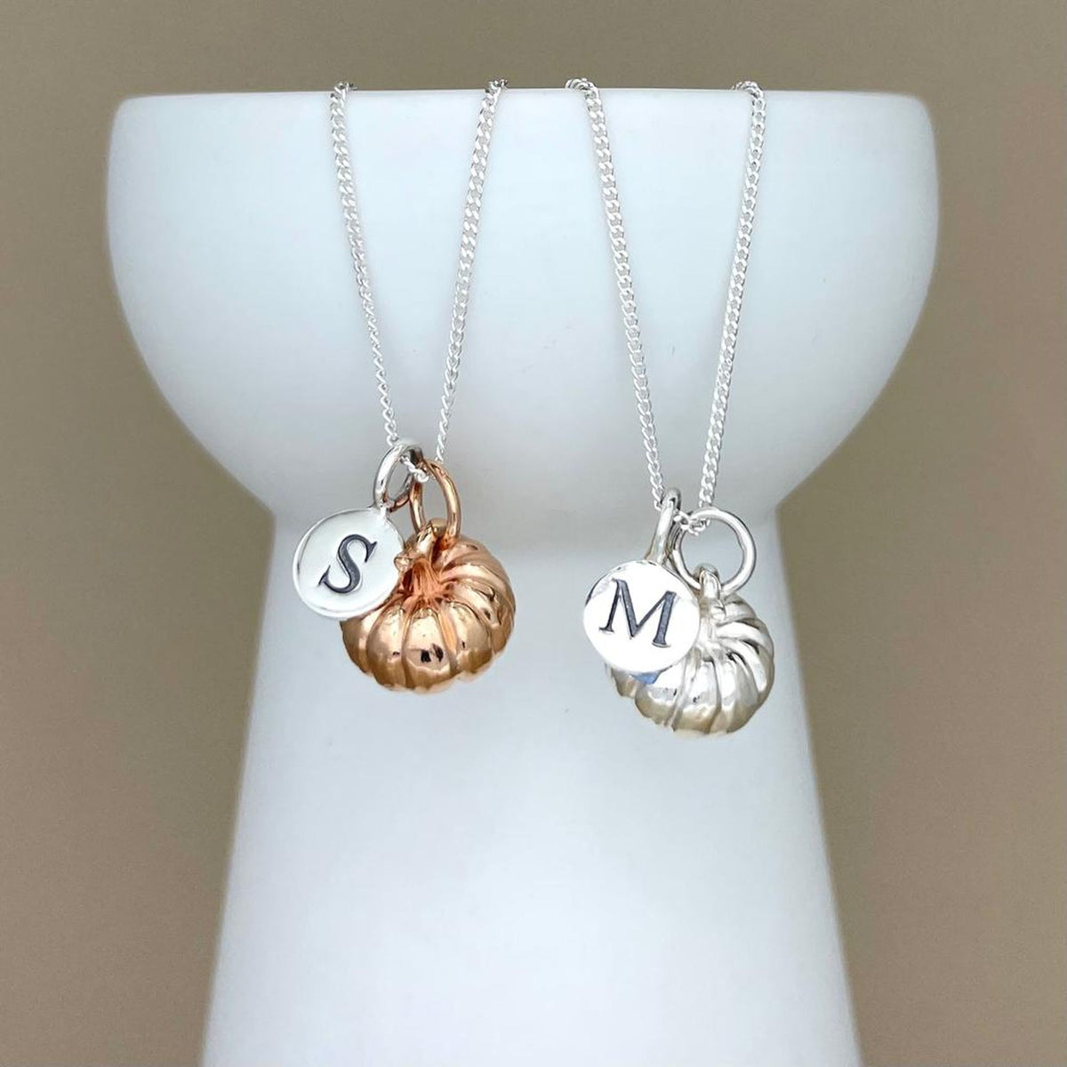 Two necklaces with initial charms on a white stand against a beige background
