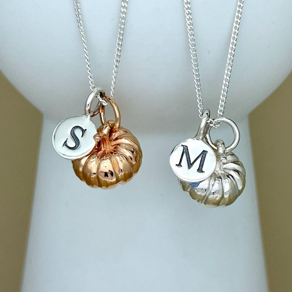 Two silver pendant necklaces displayed on a white jewelry bust. One necklace has a rose gold pumpkin charm and a silver disc charm engraved with the letter 'S'. One necklace has a silver pumpkin charm and a silver disc charm engraved with the letter 'M'.