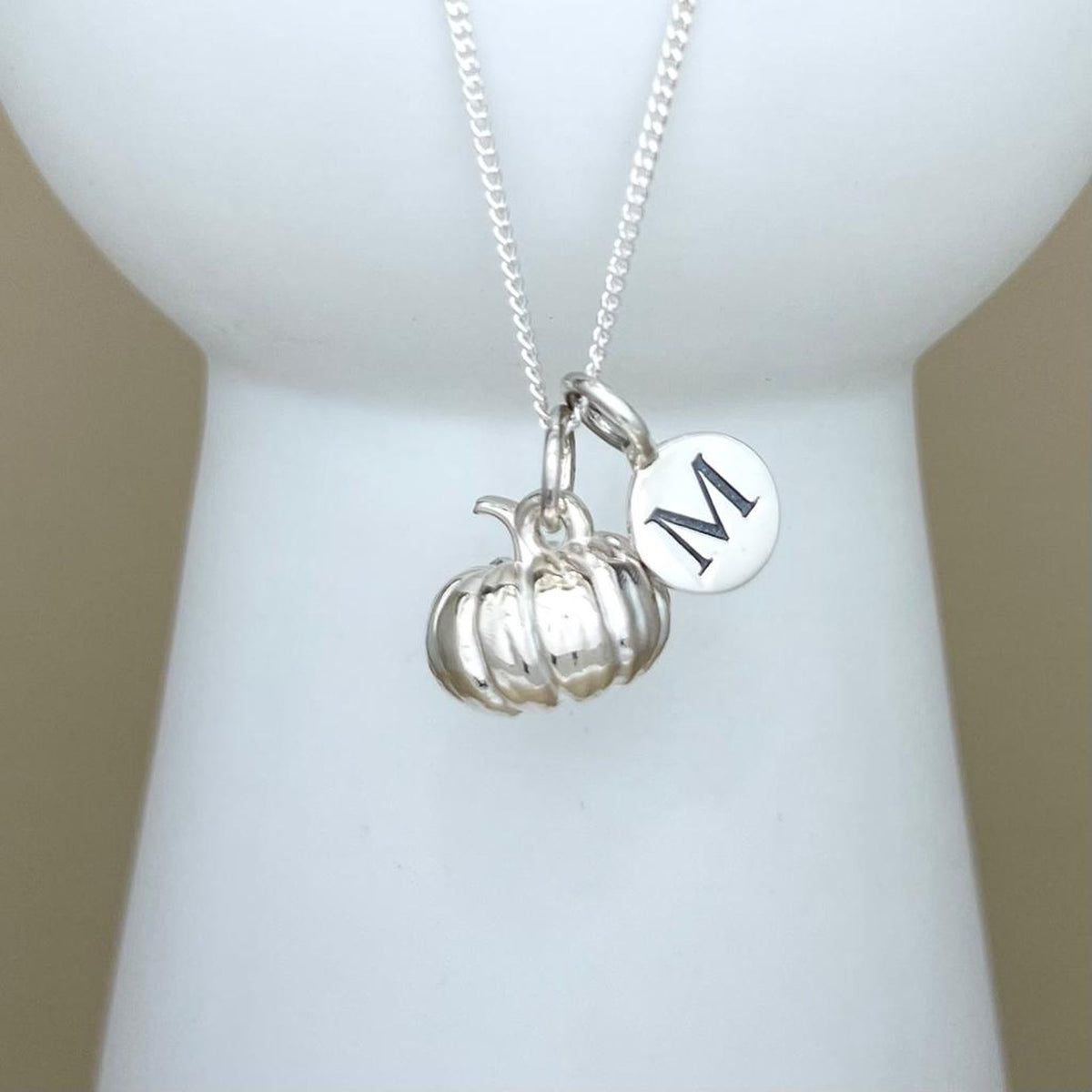 A silver necklace with a small pumpkin charm and a round initial 'M' disc charm, displayed on a white mannequin bust.