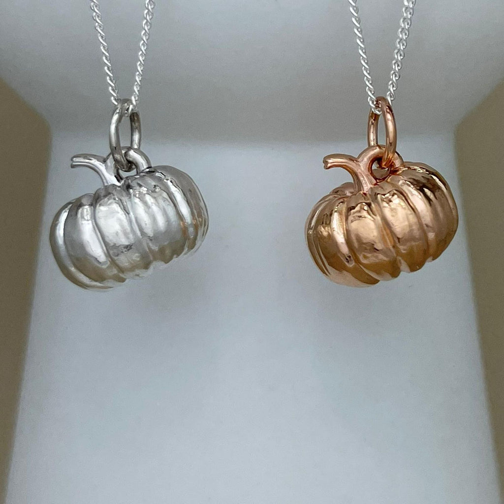Two pumpkin-shaped pendant necklaces hanging from chains, one in silver and one in rose gold, displayed against a soft white background.