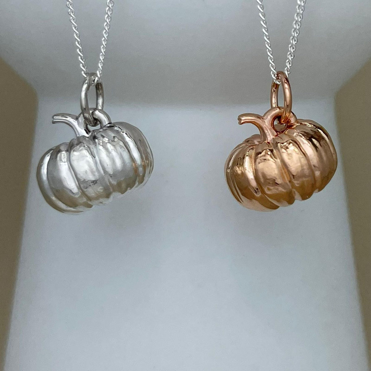 Two pumpkin-shaped pendant necklaces hanging from chains, one in silver and one in rose gold, displayed against a soft white background.