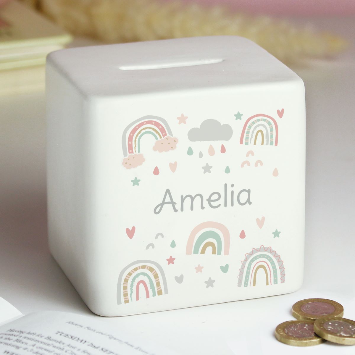 White ceramic money box with rainbow design and 'Amelia' text, placed on a surface with coins.