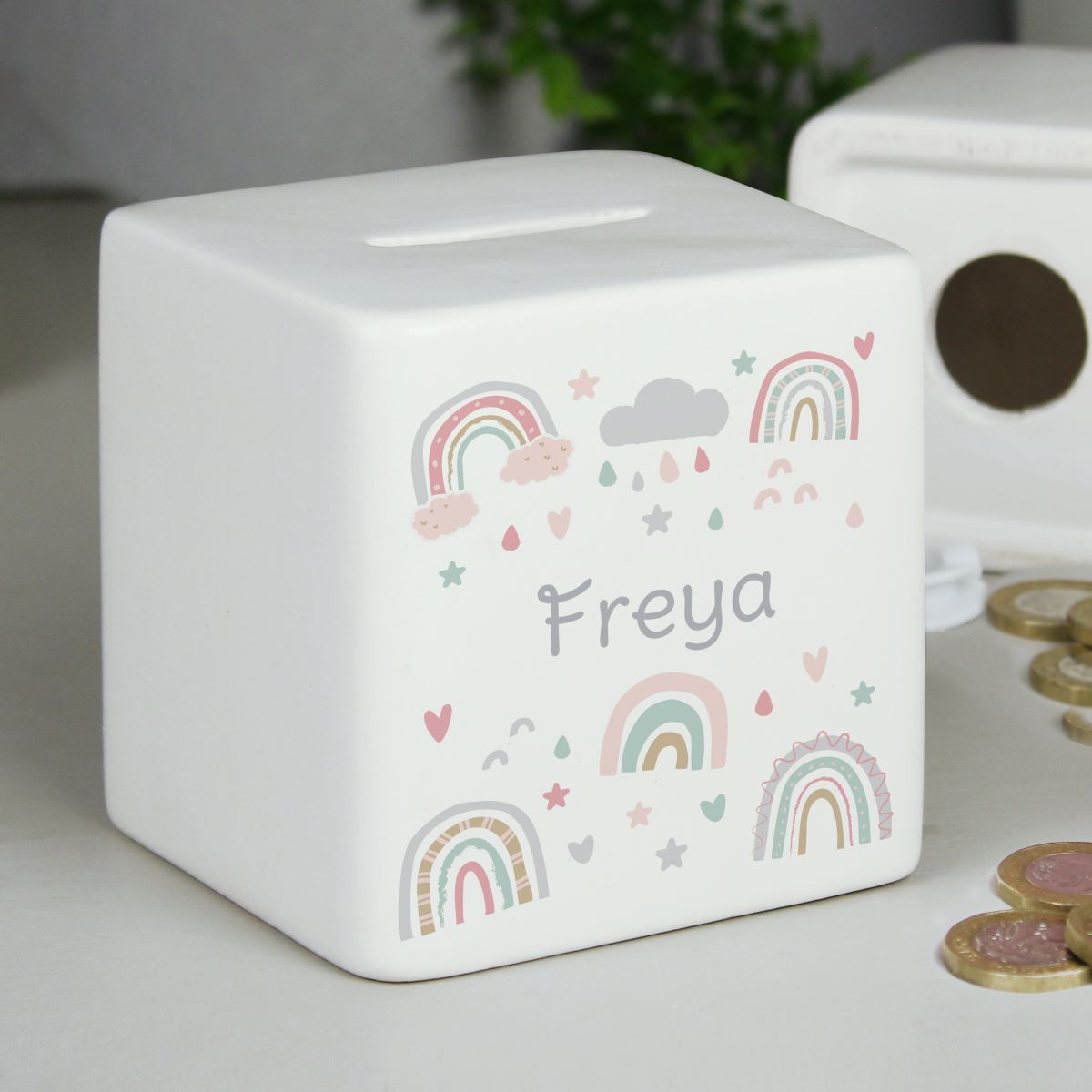 A white cube-shaped tissue box cover or money box with a slot on top, decorated with pastel rainbow illustrations, stars, hearts, and the name 'Freya' in gray lettering. It sits on a tabletop with coins nearby and a blurred plant and another box in the background.