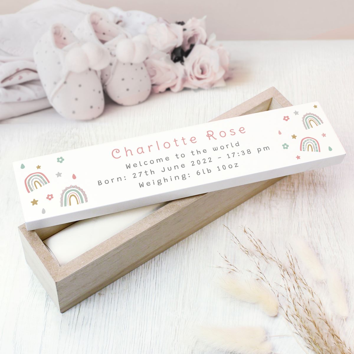 A personalized baby keepsake wooden box, with a white label printed with the name 'Charlotte Rose' and birth details, surrounded by soft baby props including pink booties and dried grass on a white surface.
