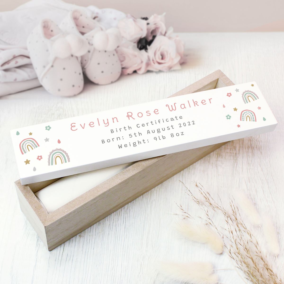 A personalized baby keepsake box containing a birth certificate, shown in a styled nursery setting with baby booties and soft pink roses in the background.
