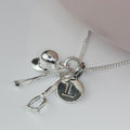 A silver necklace with a riding hat shaped charm, a small horse crop charm, a small stirrup charm, and a round initial 'I' pendant on a silver chain, displayed on a light pink surface.