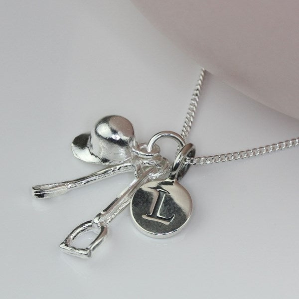 A silver necklace with a riding hat shaped charm, a small horse crop charm, a small stirrup charm, and a round initial 'I' pendant on a silver chain, displayed on a light pink surface.