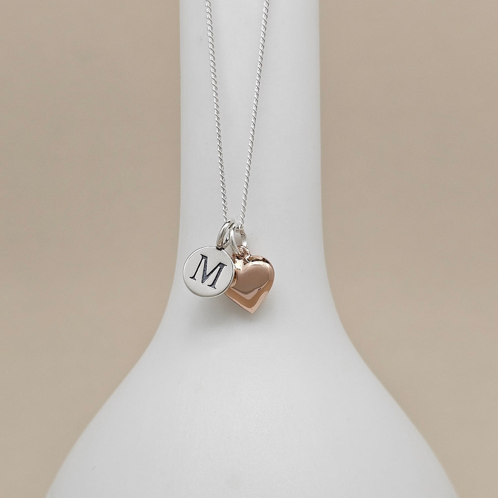 A silver chain necklace with two charms hanging from a white display bust: a round silver disc engraved with the letter M and a small rose-gold heart charm, against a soft beige background.