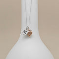 A silver chain necklace with two charms hanging from a white display bust: a round silver disc engraved with the letter M and a small rose-gold heart charm, against a soft beige background.