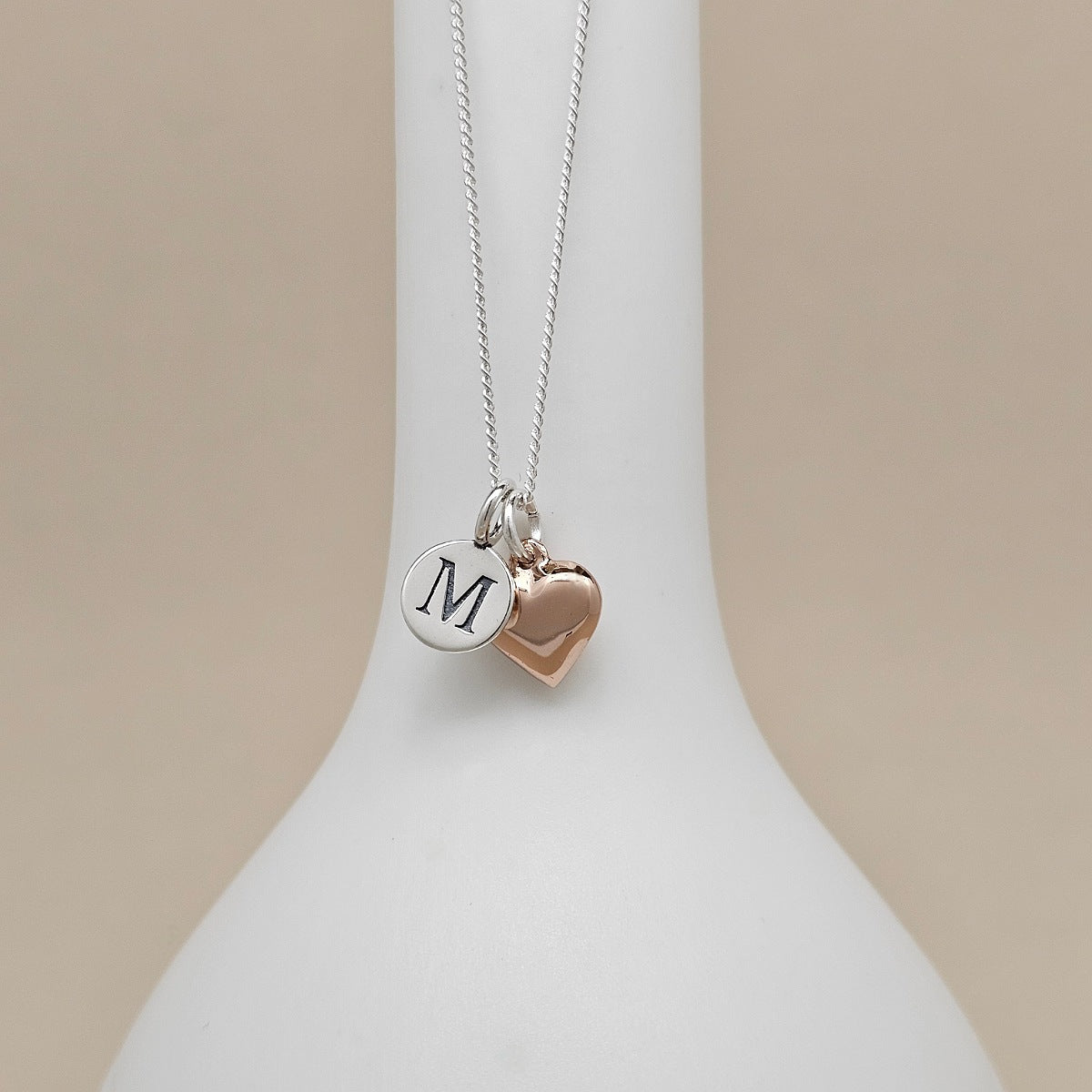 A silver chain necklace with two charms hanging from a white display bust: a round silver disc engraved with the letter M and a small rose-gold heart charm, against a soft beige background.