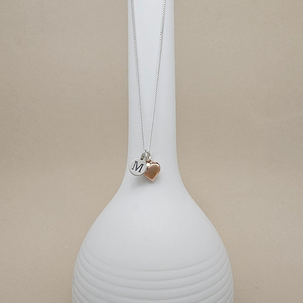 A silver chain necklace with two charms hanging from a white display bust: a round silver disc engraved with the letter M and a small rose-gold heart charm, against a soft beige background.