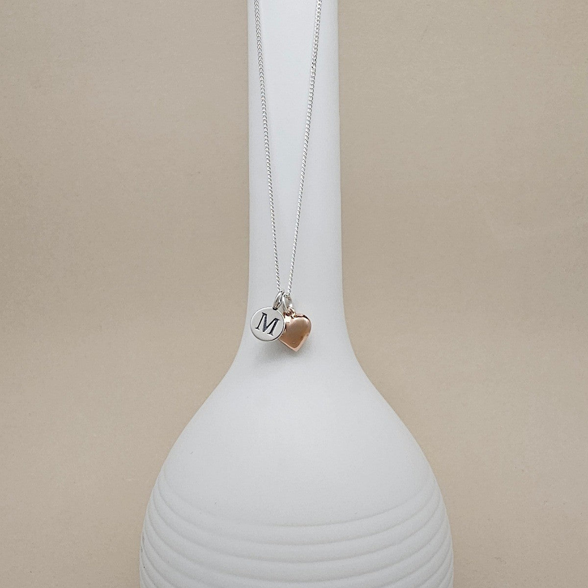 A silver chain necklace with two charms hanging from a white display bust: a round silver disc engraved with the letter M and a small rose-gold heart charm, against a soft beige background.