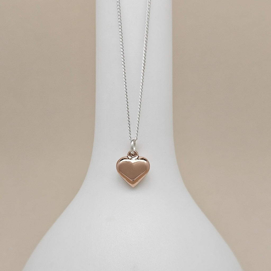 A heart-shaped pendant necklace in a warm rose-gold tone on a delicate silver chain, displayed on a white jewelry bust against a soft beige background.