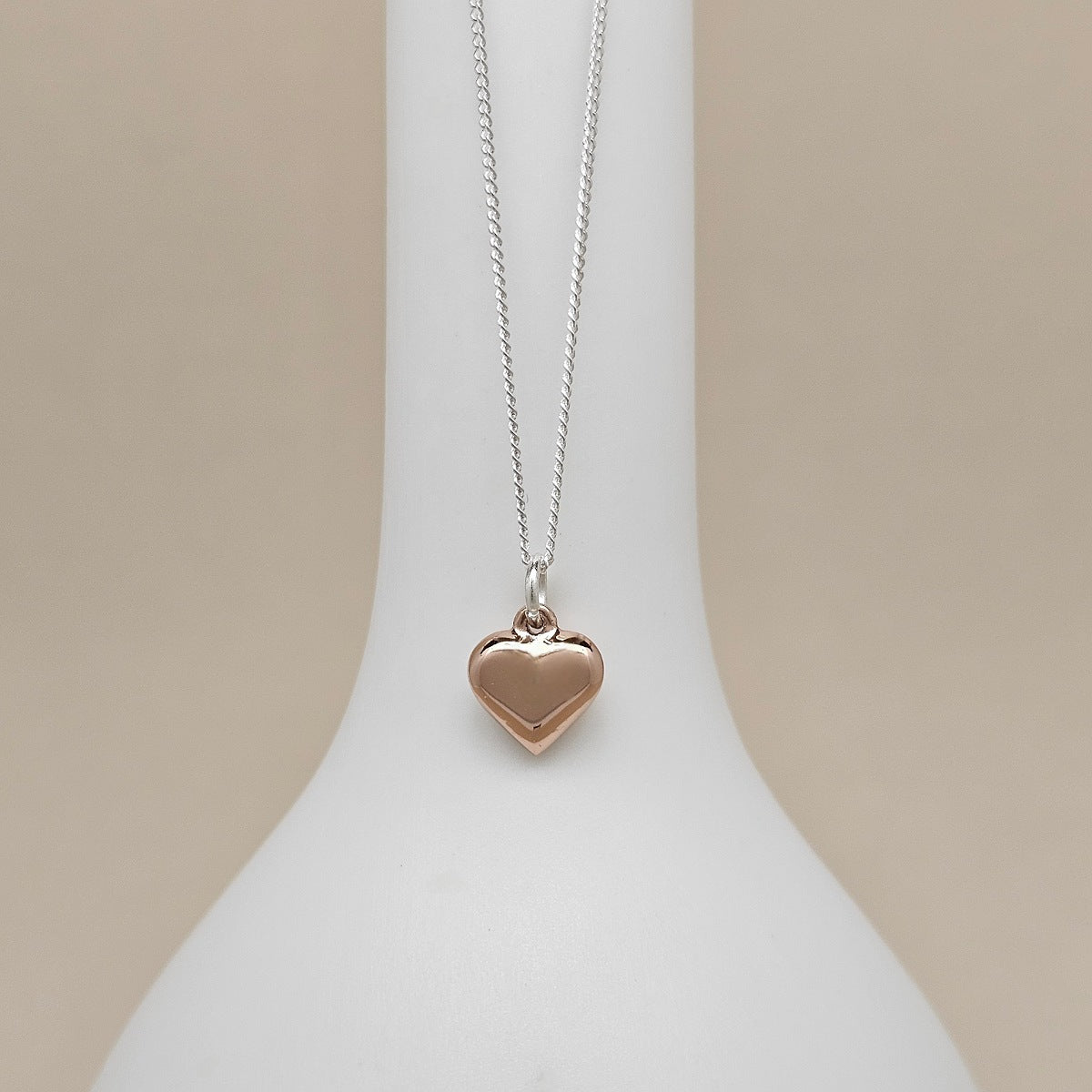 A heart-shaped pendant necklace in a warm rose-gold tone on a delicate silver chain, displayed on a white jewelry bust against a soft beige background.
