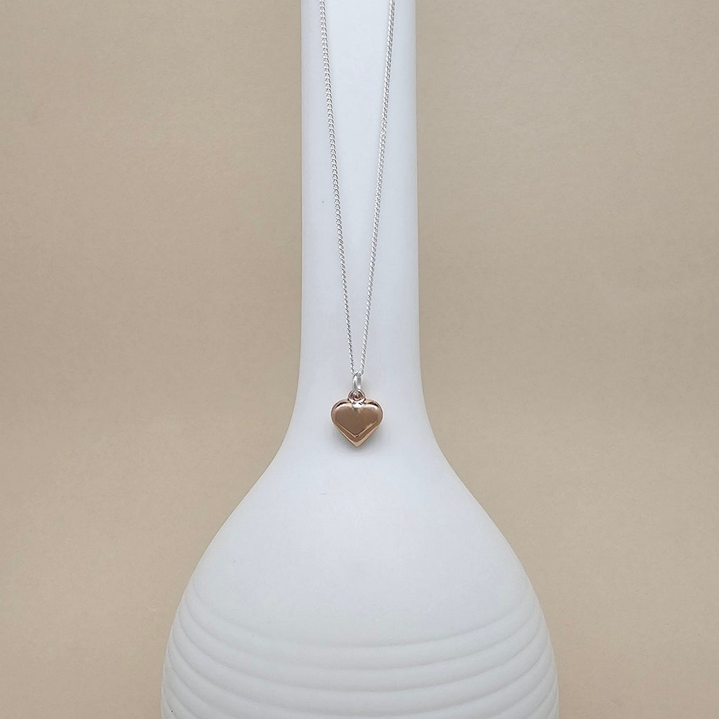 A silver chain necklace with a small polished rose gold heart pendant, displayed on a ribbed white ceramic vase against a solid beige background.