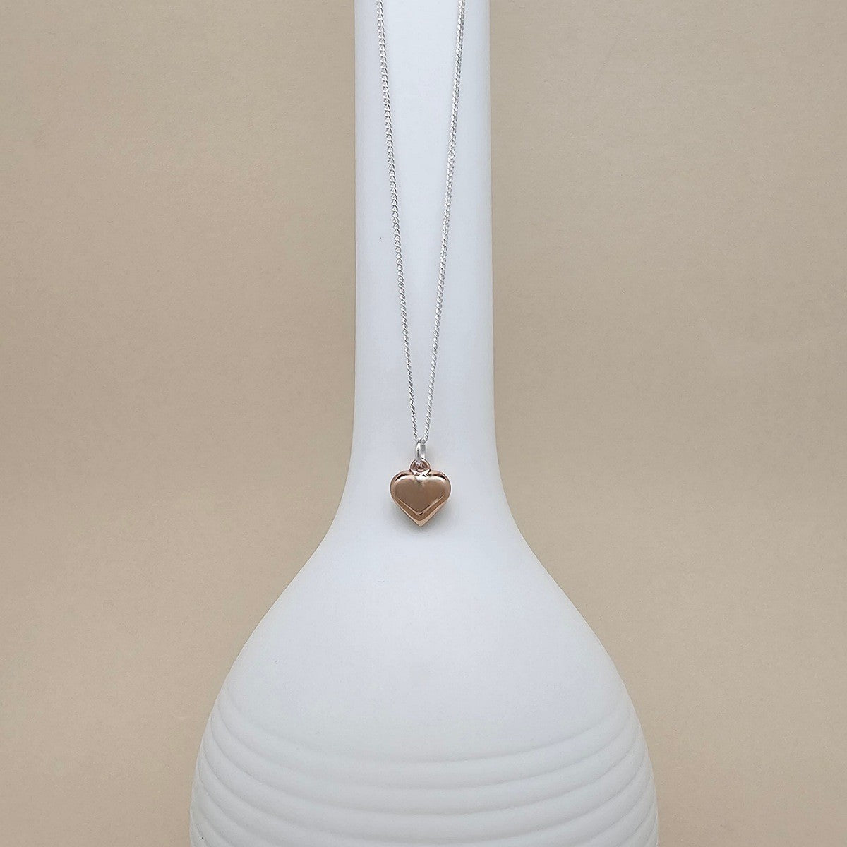 A silver chain necklace with a small polished rose gold heart pendant, displayed on a ribbed white ceramic vase against a solid beige background.