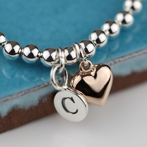 Silver beaded bracelet with a heart charm and round charm with letter 'C' on a blue and brown background.