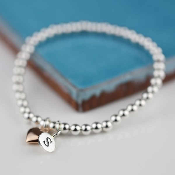 A silver beaded stretch bracelet with a small engraved silver charm and a rose gold heart charm, displayed on a white surface with a blue ceramic tile in the background.