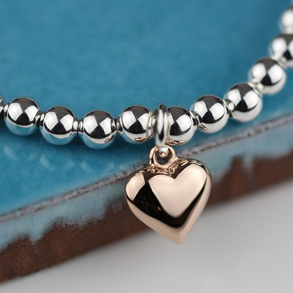 A close-up of a beaded bracelet with shiny silver-toned round beads and a polished rose gold heart charm, resting on a textured blue and brown surface.