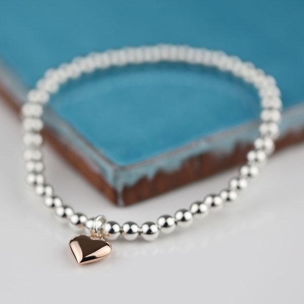 A silver beaded stretch bracelet with a small rose gold heart charm, displayed on a blue textured card over a light gray surface.