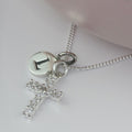 A silver initial necklace featuring a cross charm adorned with clear rhinestones and a round disc charm engraved with the letter 'I', displayed on a soft pink surface.