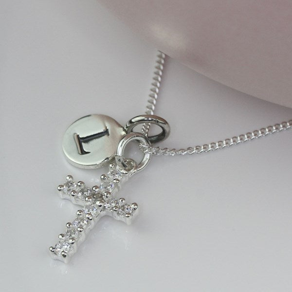 A silver initial necklace featuring a cross charm adorned with clear rhinestones and a round disc charm engraved with the letter 'I', displayed on a soft pink surface.