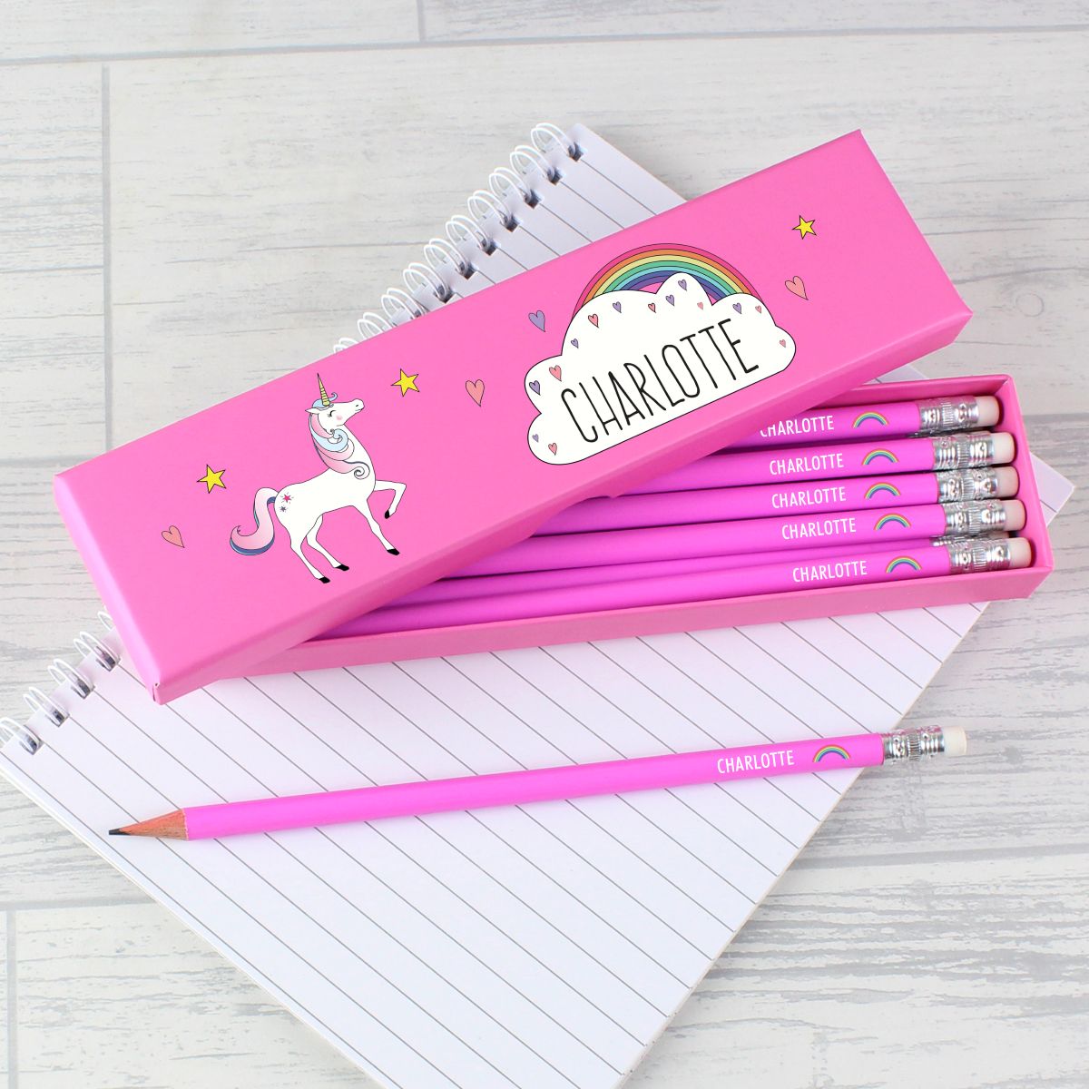 A pink personalized pencil case with a unicorn illustration and the name 'CHARLOTTE' on the lid, shown open with several matching pink pencils inside. A loose pink pencil lies in front on top of lined notebook pages over a light wood surface.