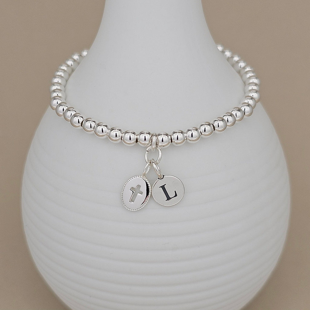 A silver beaded bracelet with a cross charm and letter 'L' charm, displayed on a white ribbed ceramic vase against a warm beige background.