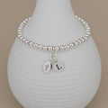 A silver beaded bracelet with a cross charm and letter 'L' charm, displayed on a white ribbed ceramic vase against a warm beige background.