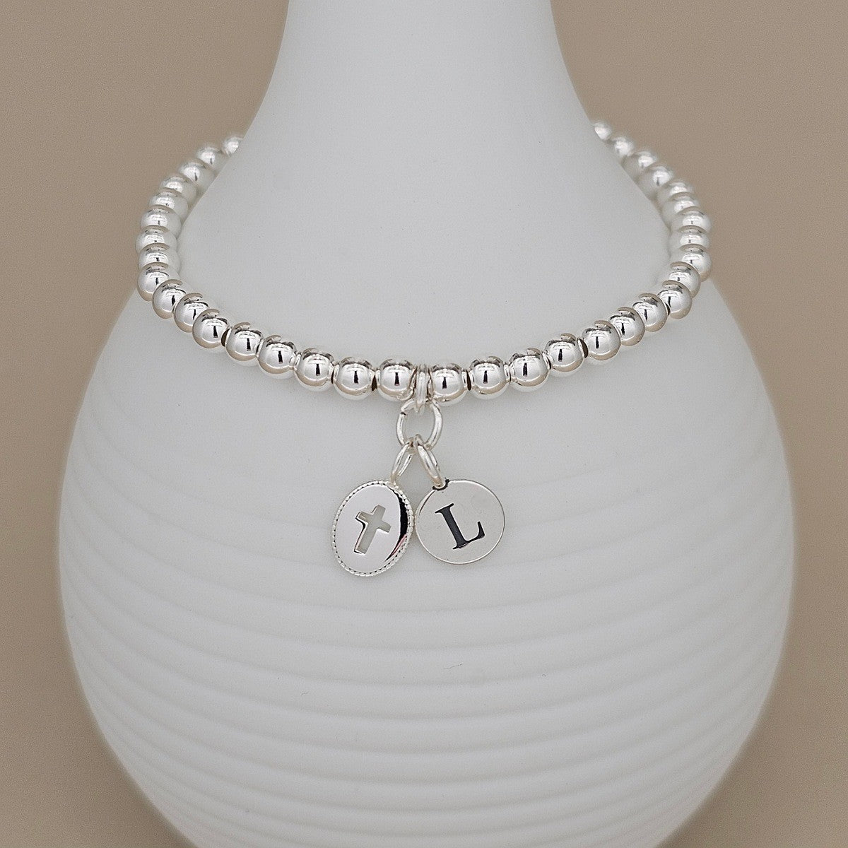 A silver beaded bracelet with a cross charm and letter 'L' charm, displayed on a white ribbed ceramic vase against a warm beige background.