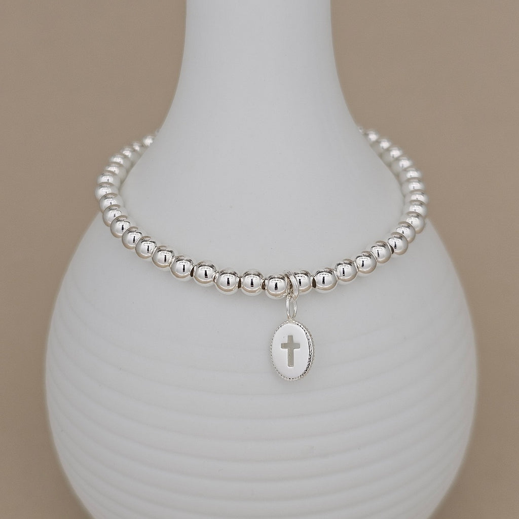 A silver beaded bracelet with an oval cross charm, displayed on a white ribbed ceramic vase against a neutral beige background.