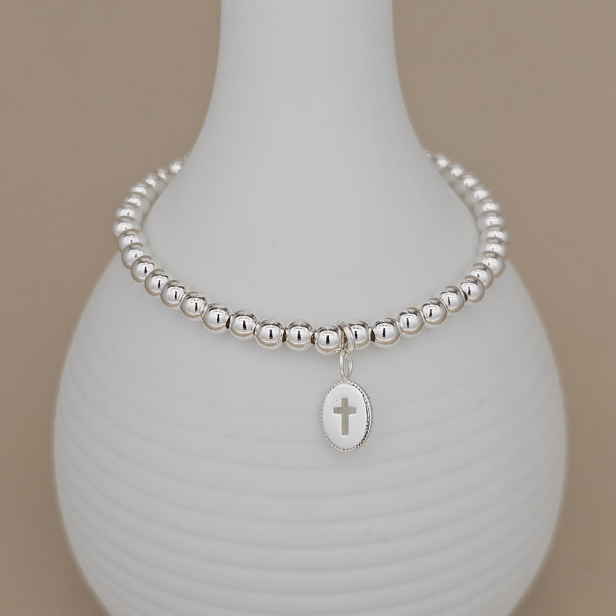 A silver beaded bracelet with an oval cross charm, displayed on a white ribbed ceramic vase against a neutral beige background.
