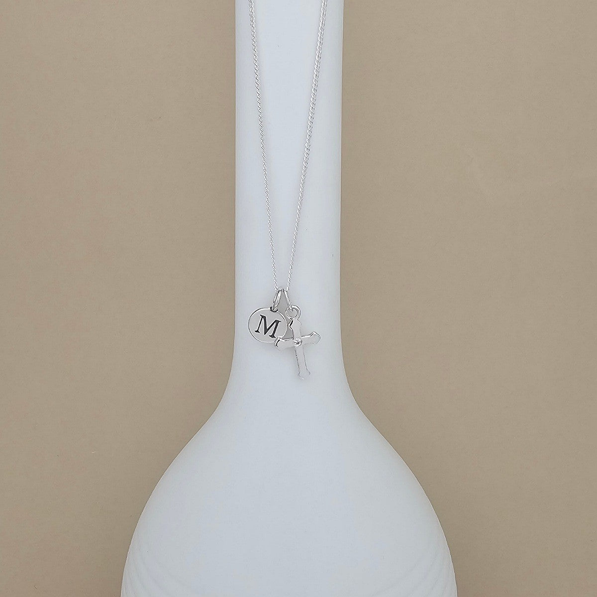 A silver necklace with a small cross pendant and a charm engraved with the letter 'M', displayed on a white vase against a beige background.