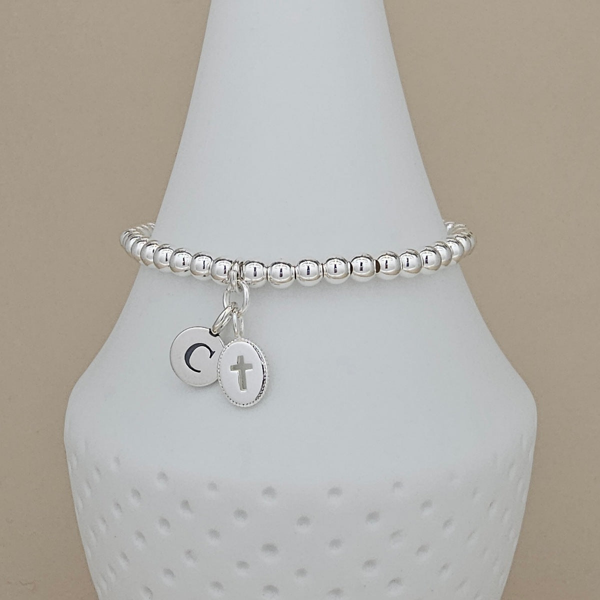 A silver beaded bracelet with two dangling charms, one engraved with a letter C and the other with a cross, displayed on a white cone-shaped jewelry stand against a beige background.