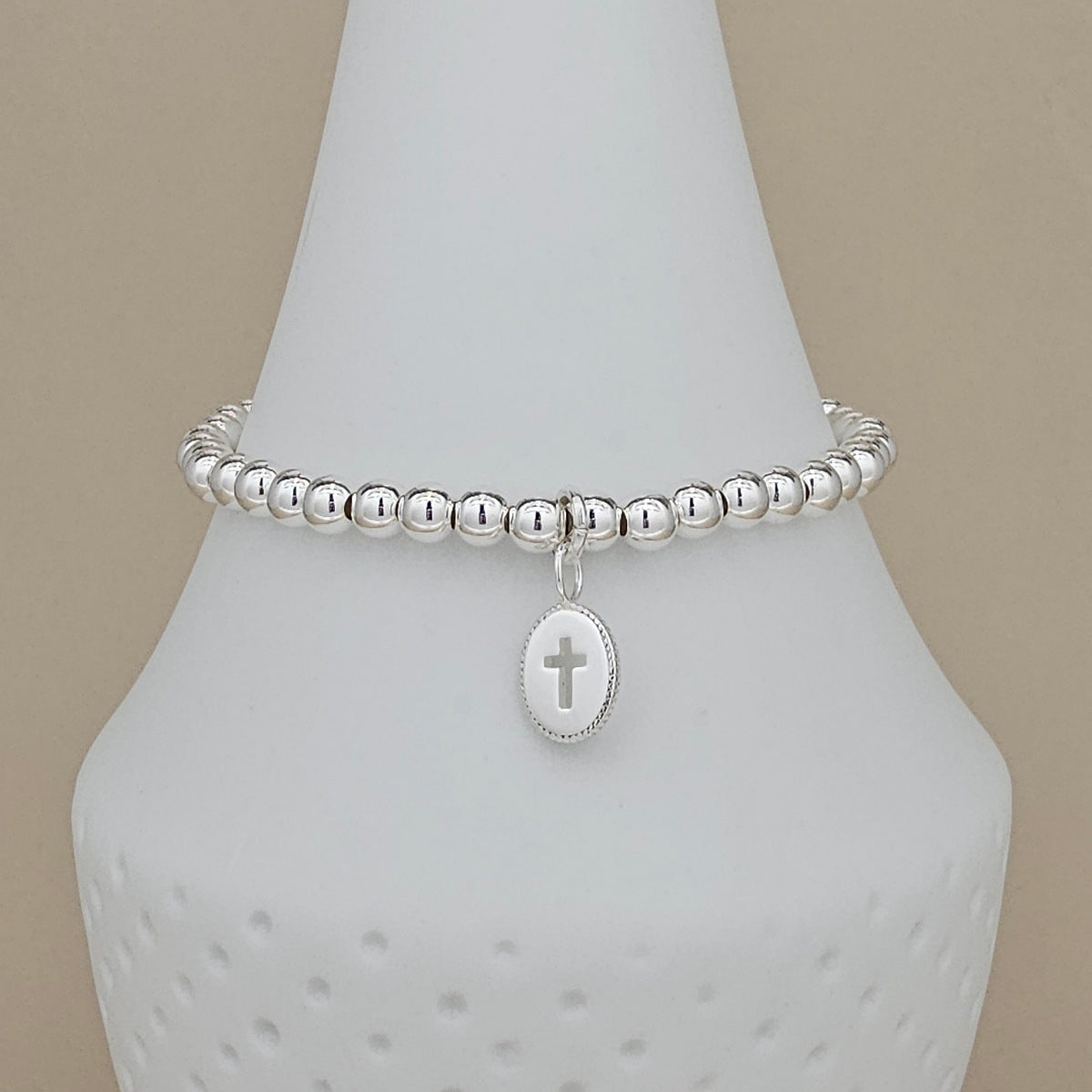 A silver beaded bracelet with a small oval pendant featuring a cross, displayed on a white cone-shaped jewelry stand against a beige background.