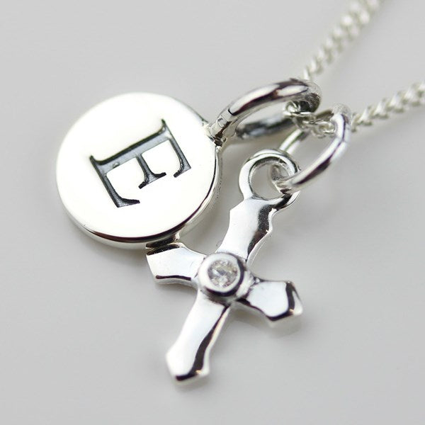A silver necklace featuring a round disc pendant engraved with the letter 'H' and a polished cross charm with a small clear stone at the center, shown on a light gray background.