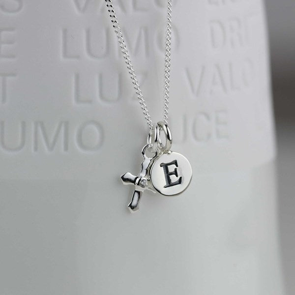 A silver necklace with a small cross charm and a round disc charm engraved with the letter 'E', hanging in front of a white textured background.