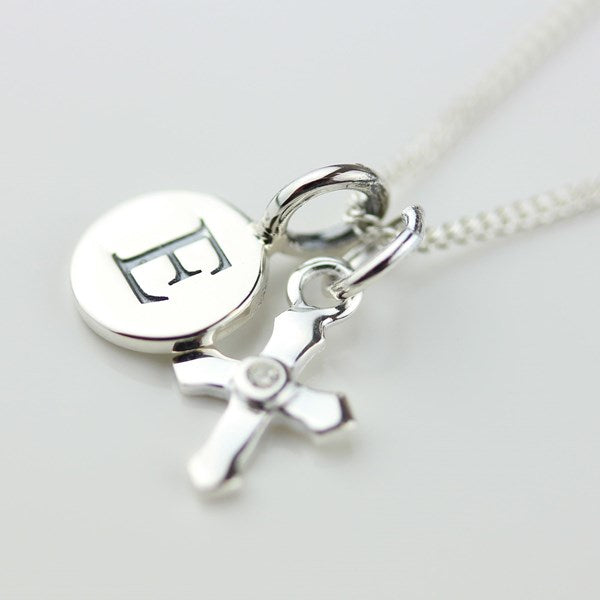 A silver necklace with a round disc charm engraved with a letter and a small cross charm, shown on a light gray background.