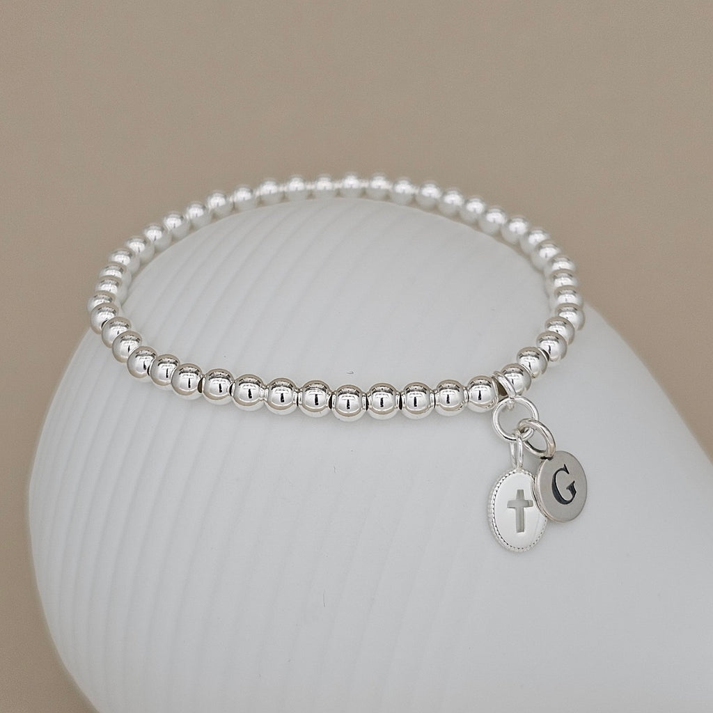 Silver beaded bracelet with cross and initial charm on a white surface